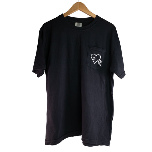 Polera negra Make Noise QMMG con un corazón estampado en blanco sobre el bolsillo del pecho, colgada en una percha