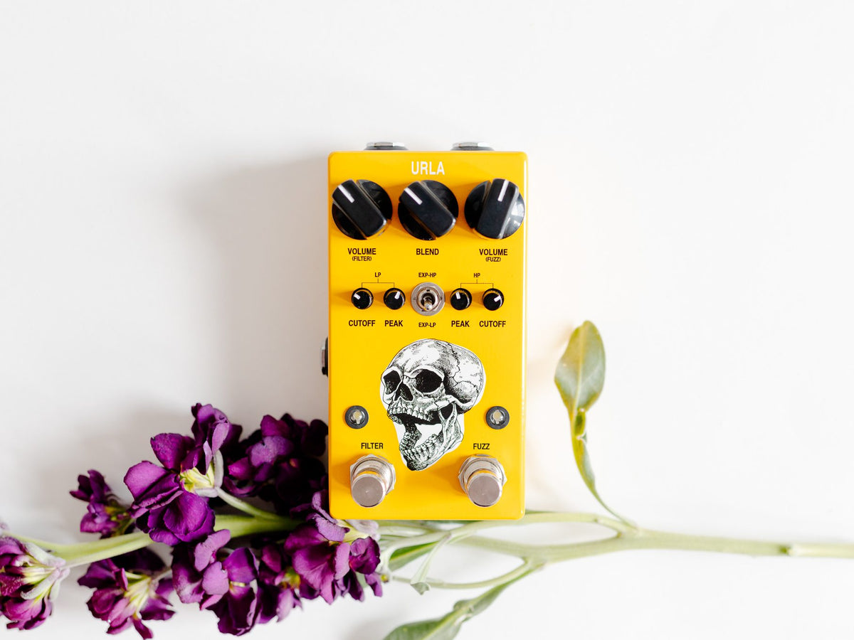 Pedal AC Noises Urla, amarillo con una calavera ilustrada, junto a flores moradas