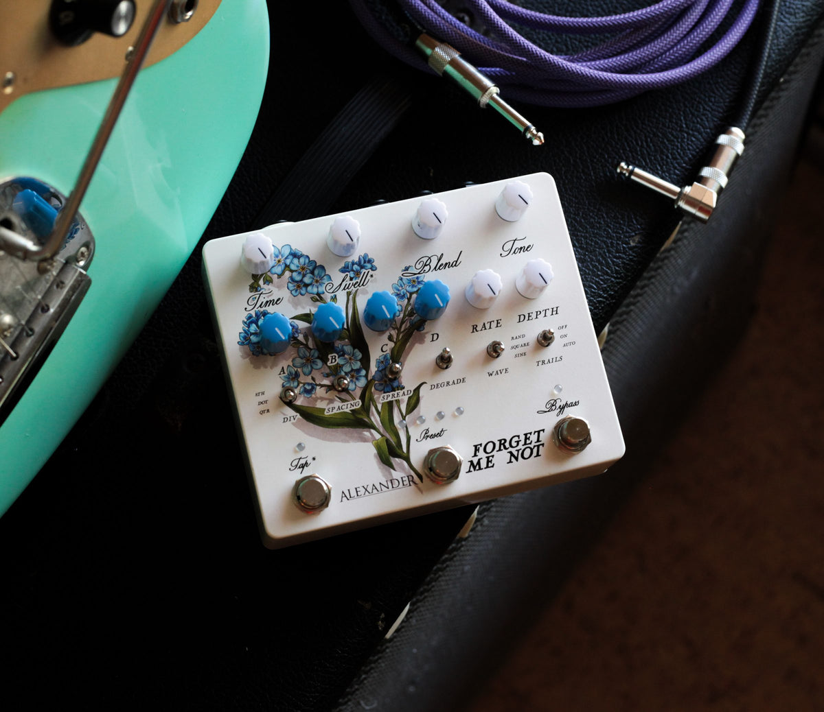 Pedal Alexander Forget-Me-Not, blanco con flores azules ilustradas, junto a una guitarra verde agua y un cable morado